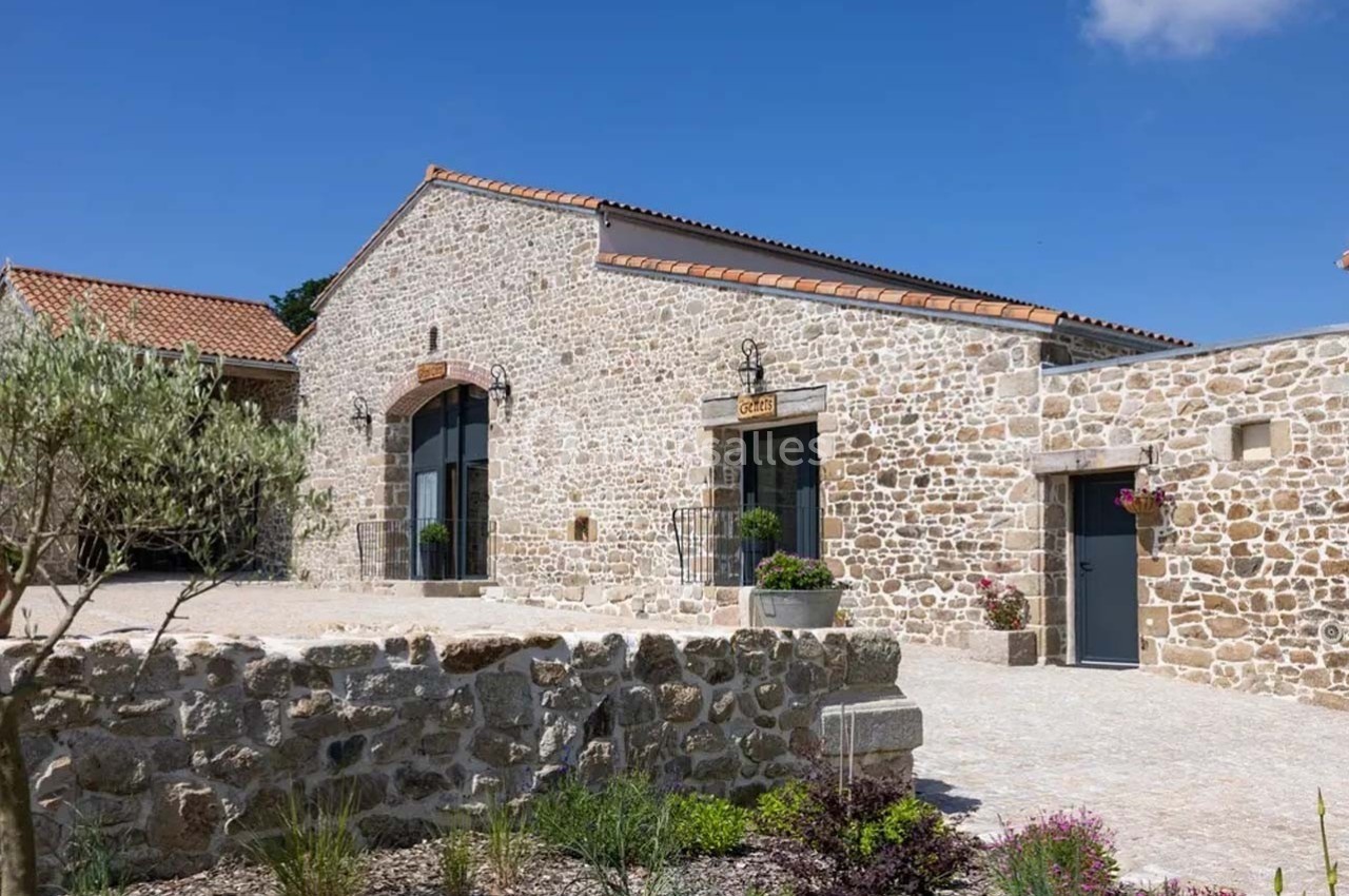 Bâtiment en pierre rénové avec toit en tuiles, grandes ouvertures vitrées et cour aménagée avec végétation.