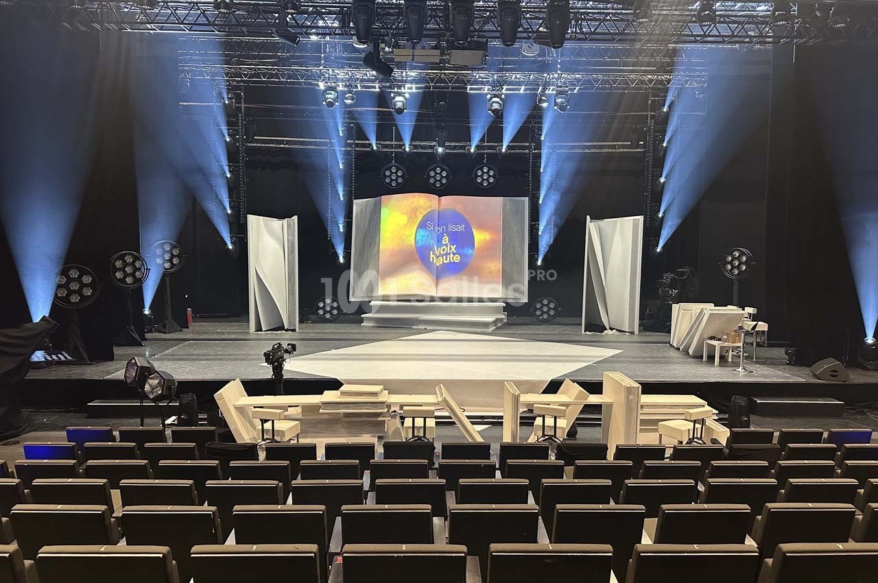Scène de spectacle avec écran central illuminé, sièges vides au premier plan et éclairages de scène activés.