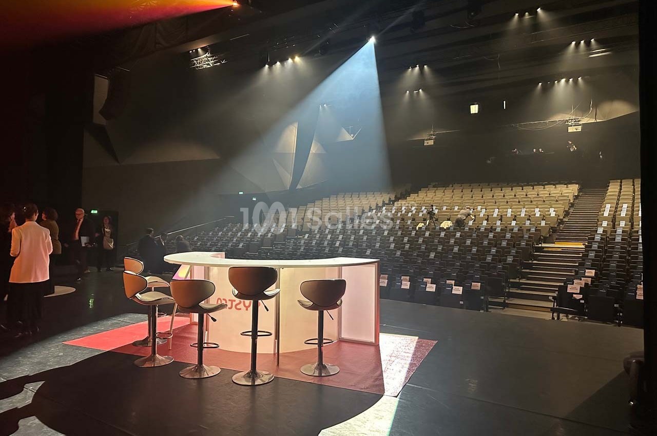 Salle de spectacle vide avec des rangées de sièges, un plateau éclairé et des tabourets devant une table blanche.