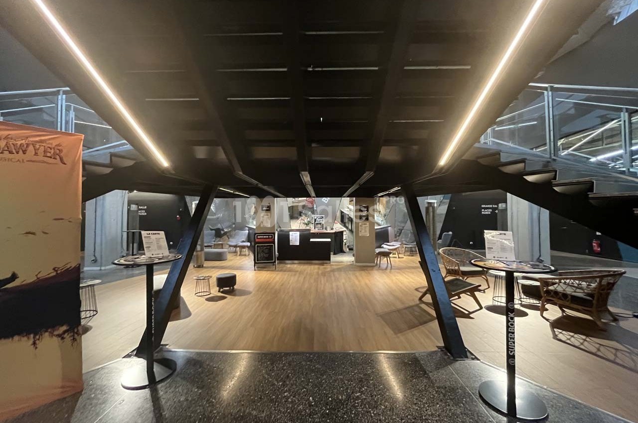 Vue d'un espace d'accueil moderne avec un escalier central, des tables hautes et des fauteuils en rotin.
