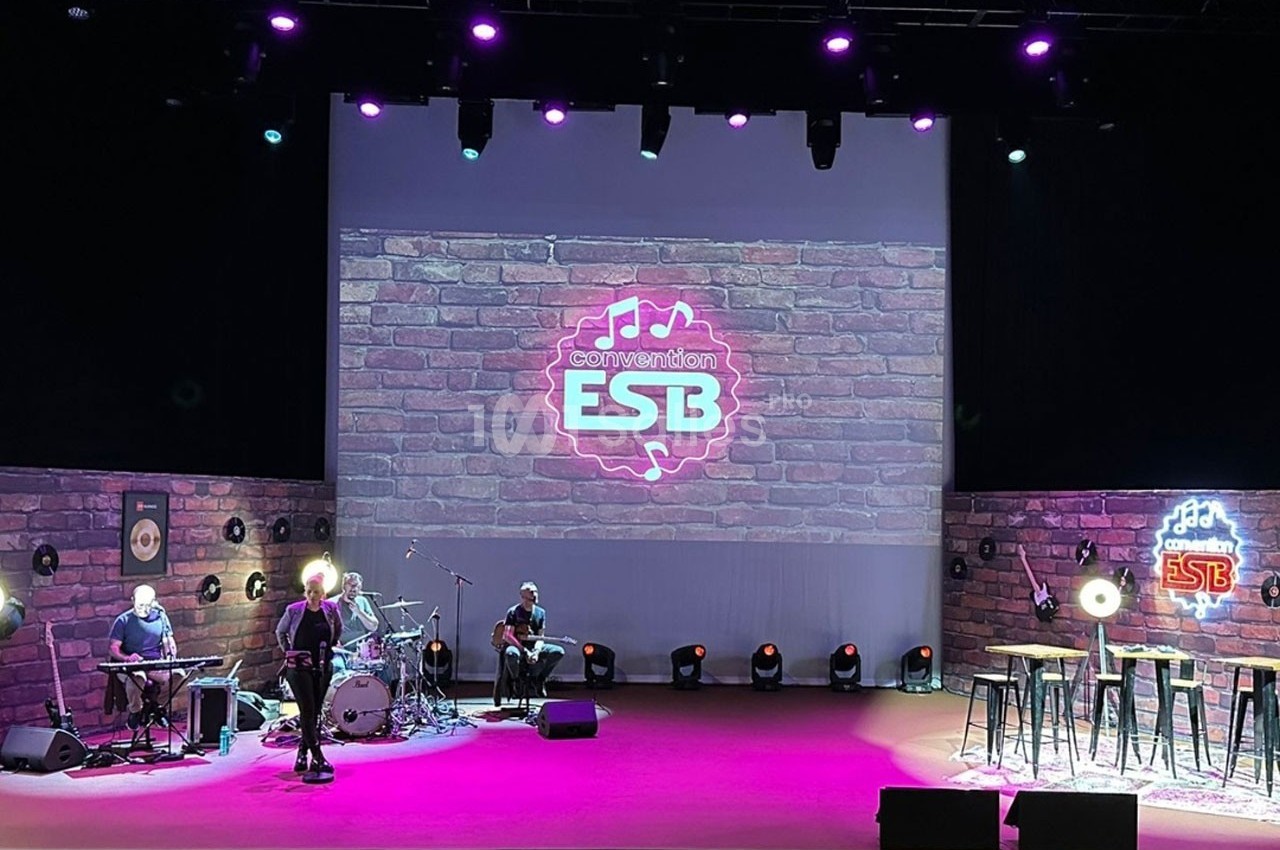 Scène éclairée avec instruments de musique, tabourets et écran affichant un logo ’ESB’ sur un fond de briques.