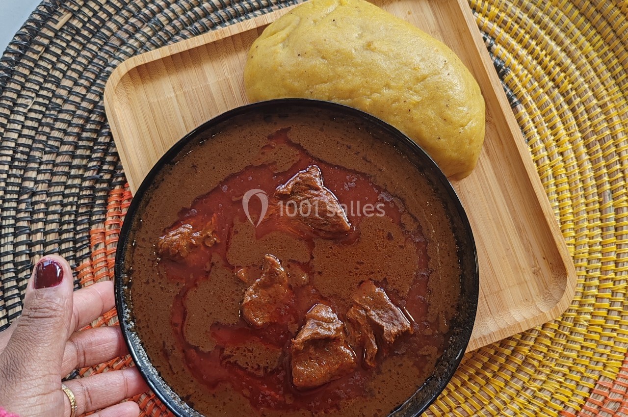 Assiette de ragoût de viande rouge dans un bol noir, accompagnée de foufou sur un plateau en bois.