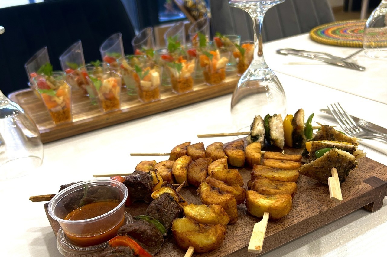 Plateau de brochettes variées avec plantains frits et sauce, accompagné de verrines colorées en arrière-plan.