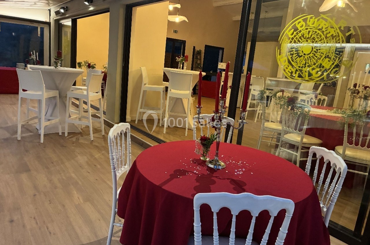 Salle décorée avec des tables rondes et hautes, nappes rouges, chaises blanches et chandeliers, ambiance élégante.