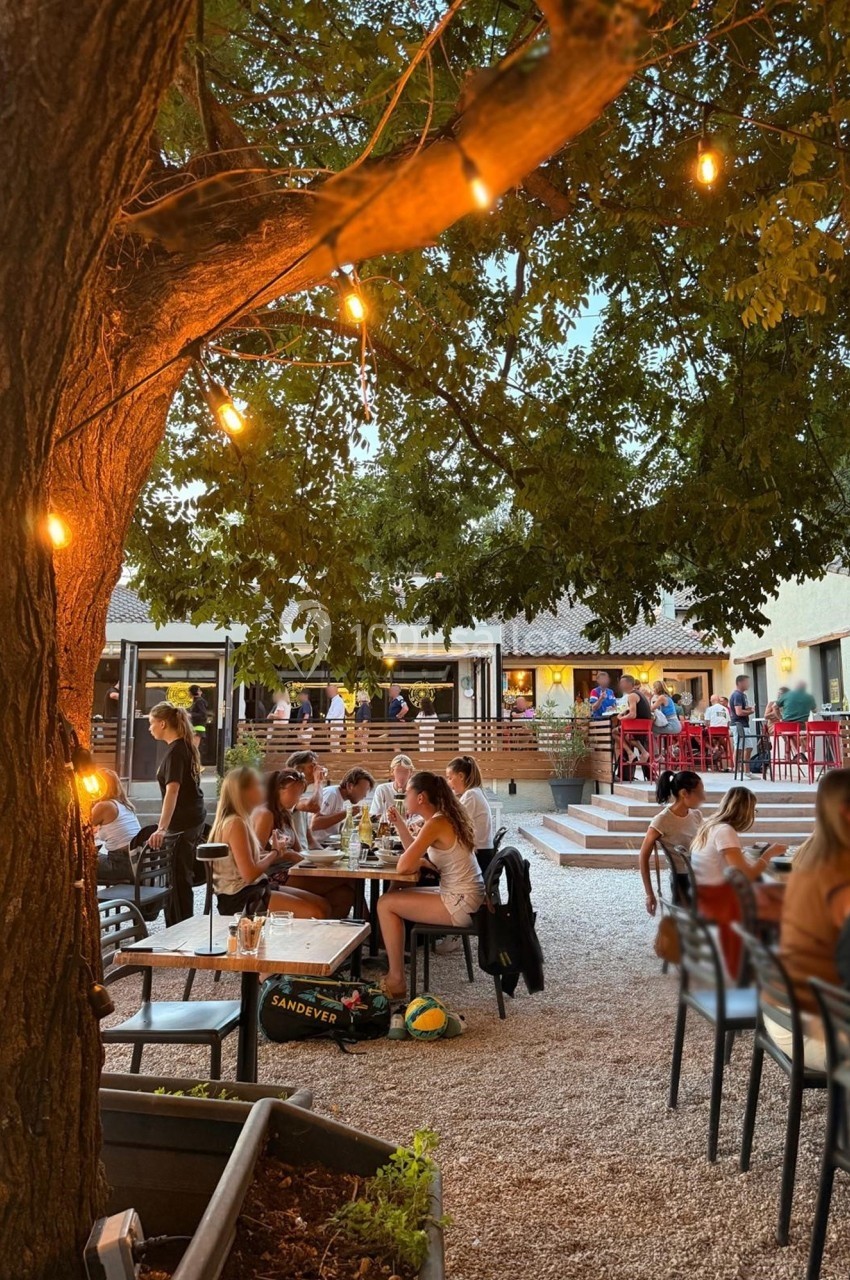 Terrasse animée d'un restaurant en soirée, avec des guirlandes lumineuses et des convives attablés sous un arbre.
