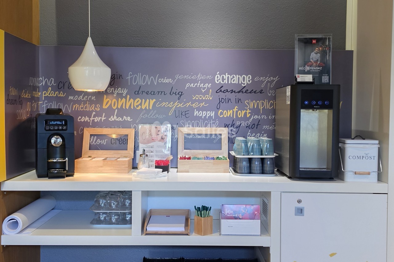 Espace d'accueil avec distributeur de café, mobilier de rangement blanc et mur orné de messages inspirants en plusieurs langues.