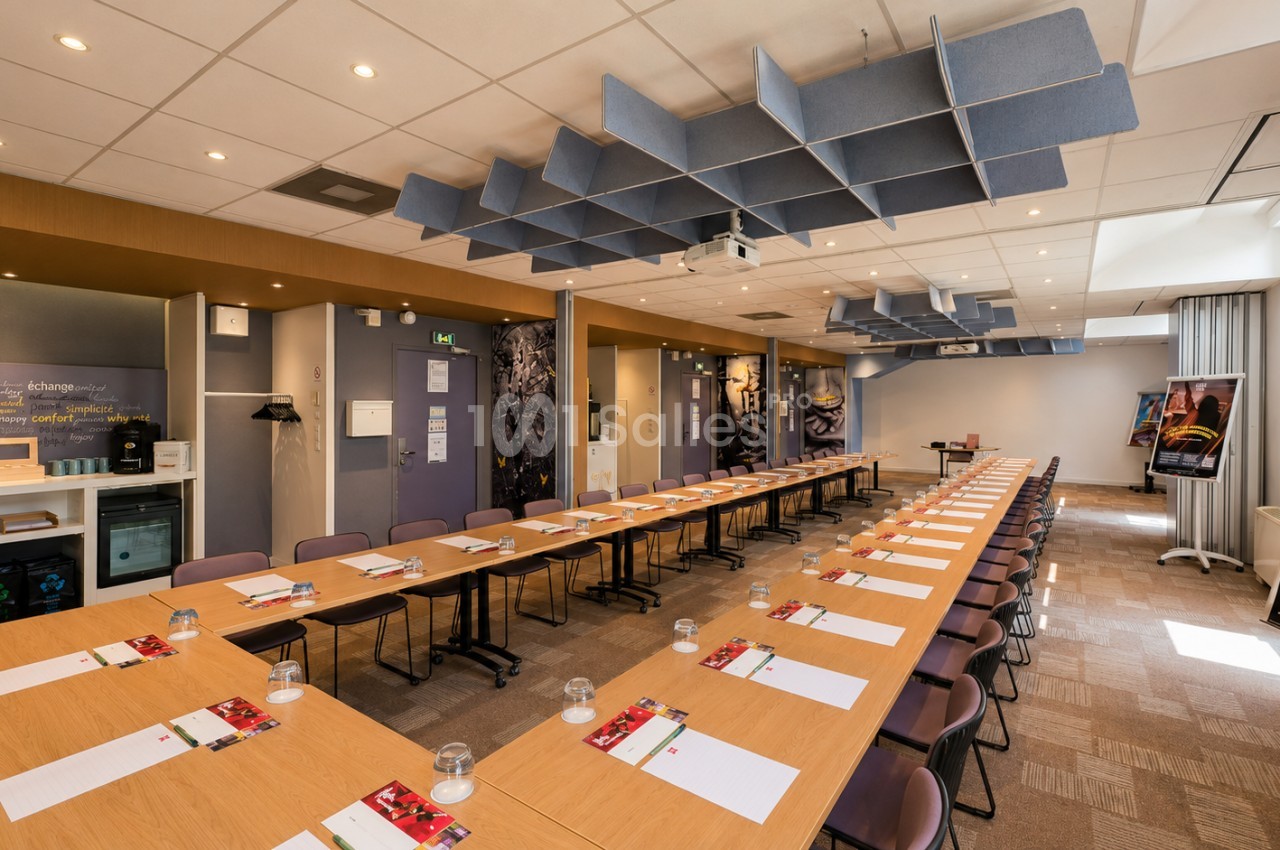 Salle de réunion avec deux tables rectangulaires en bois dressées en U, chaises marron alignées, panneaux acoustiques bleus au plafond, éclairage encastré.