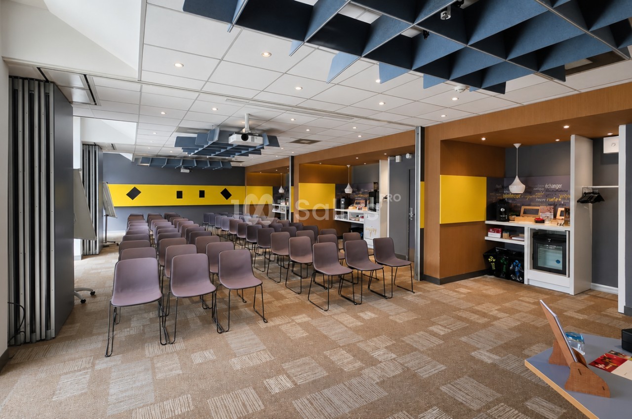 Salle de conférence aménagée avec chaises roses alignées, plafond bleu et blanc, murs ocre et jaune vif, espace ouvert sur coin café.