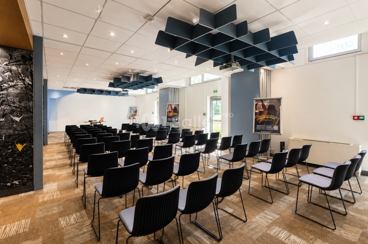 Salle de conférence avec chaises noires disposées face à un podium, plafond blanc et panneaux acoustiques bleu foncé.