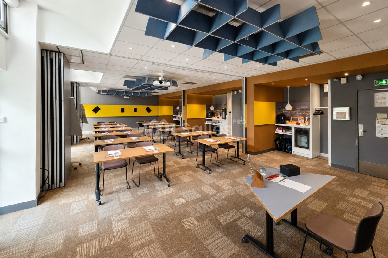 Salle de réunion modulable avec tables mobiles, chaises noires, panneaux acoustiques bleus au plafond et murs jaunes.