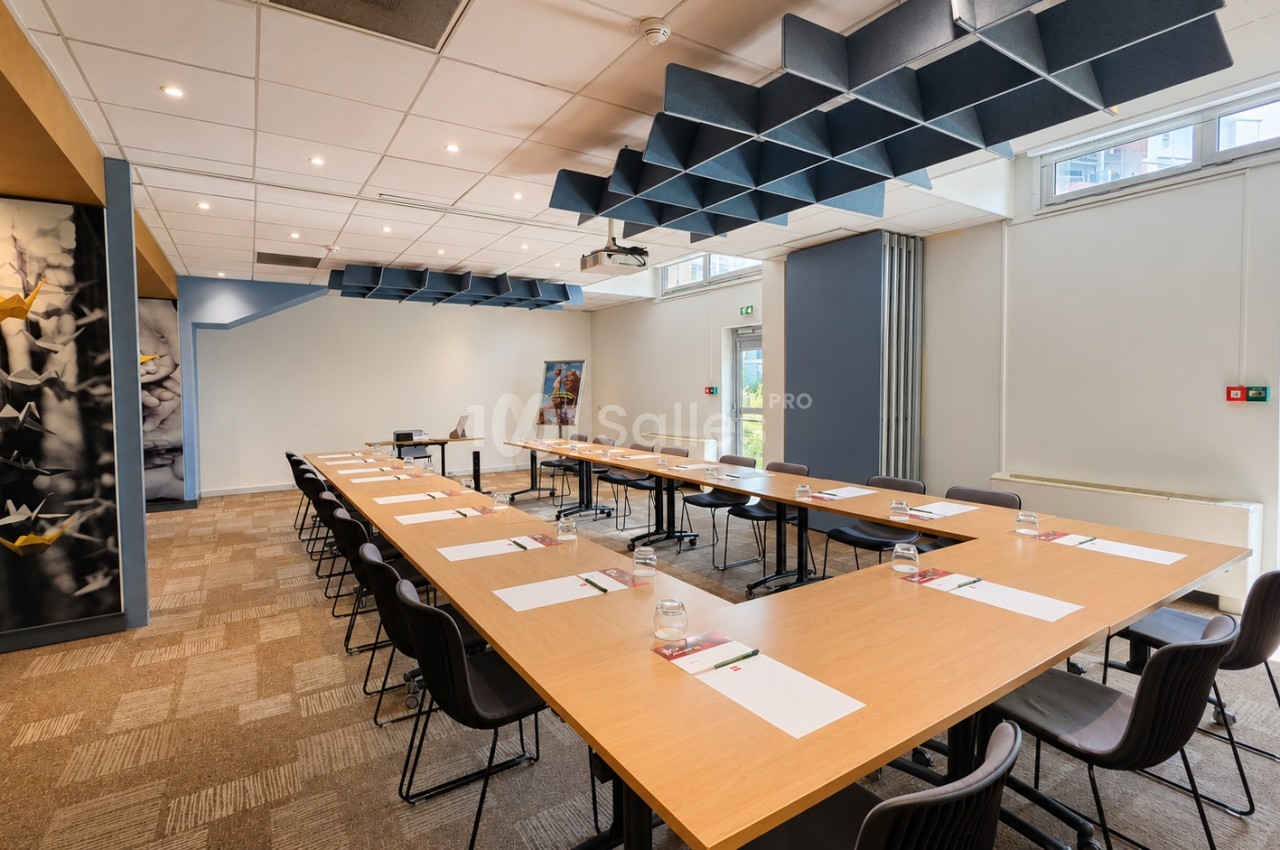 Salle de réunion avec tables en U en bois clair, chaises noires, éclairage spots et panneaux acoustiques bleus au plafond.