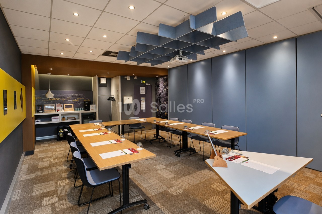 Salle de réunion modulable avec tables en bois, chaises bleues et grises, panneaux acoustiques au plafond, murs bleus et jaunes, kitchenette au fond.