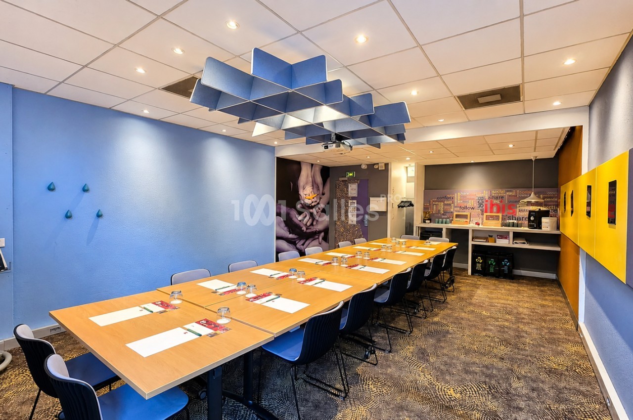 Salle de réunion avec tables en bois accueillant environ 20 places - murs bleu et jaune vif - plafonnier géométrique bleu - spots d'éclairage.