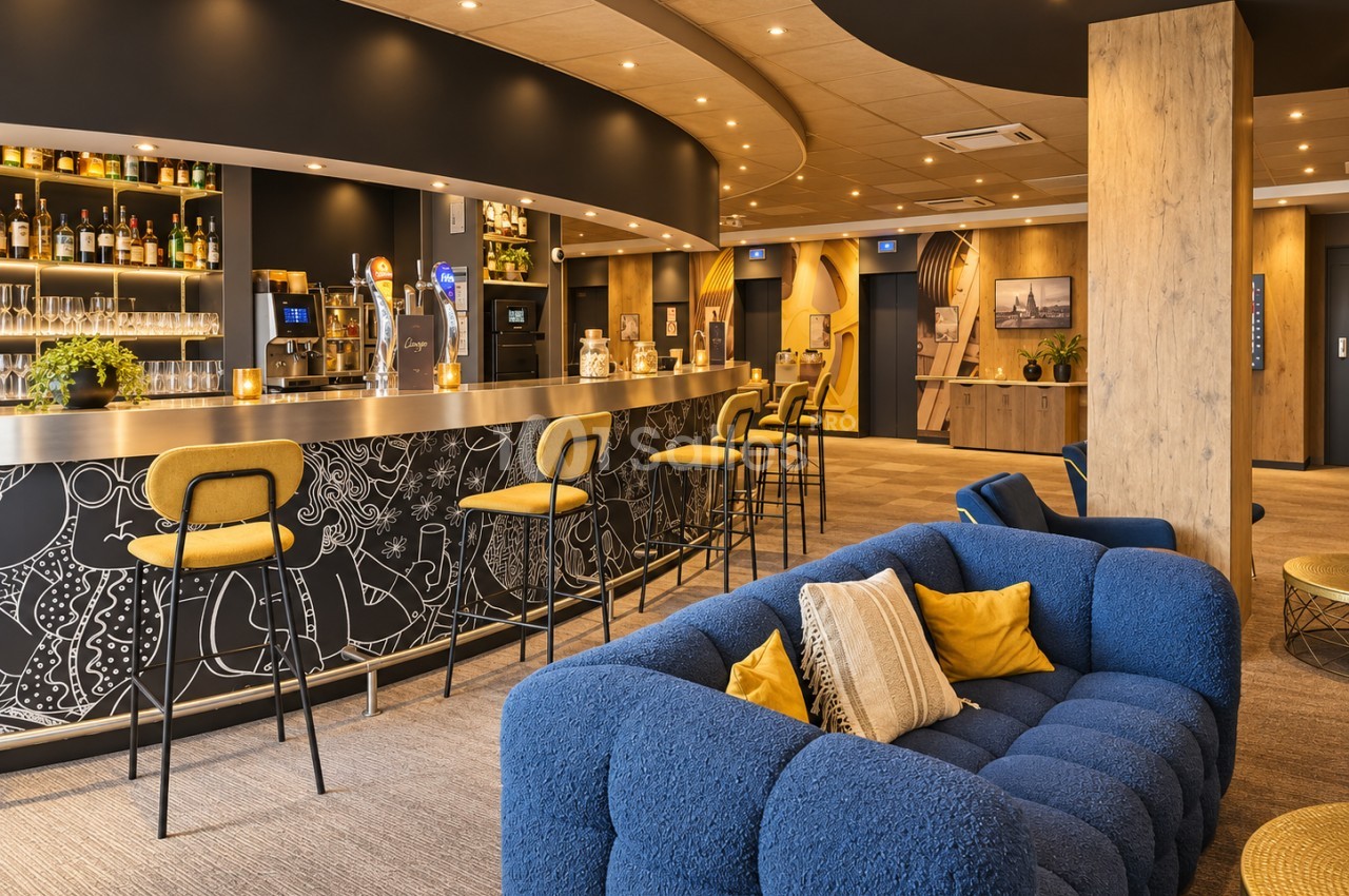 Salle intérieure avec bar équipé, chaises hautes jaunes, canapé bleu et mobilier contemporain - éclairage chaleureux.