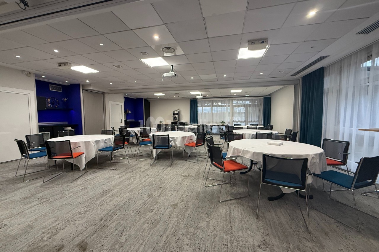Salle de réunion lumineuse avec tables rondes dressées de nappes blanches et chaises colorées.