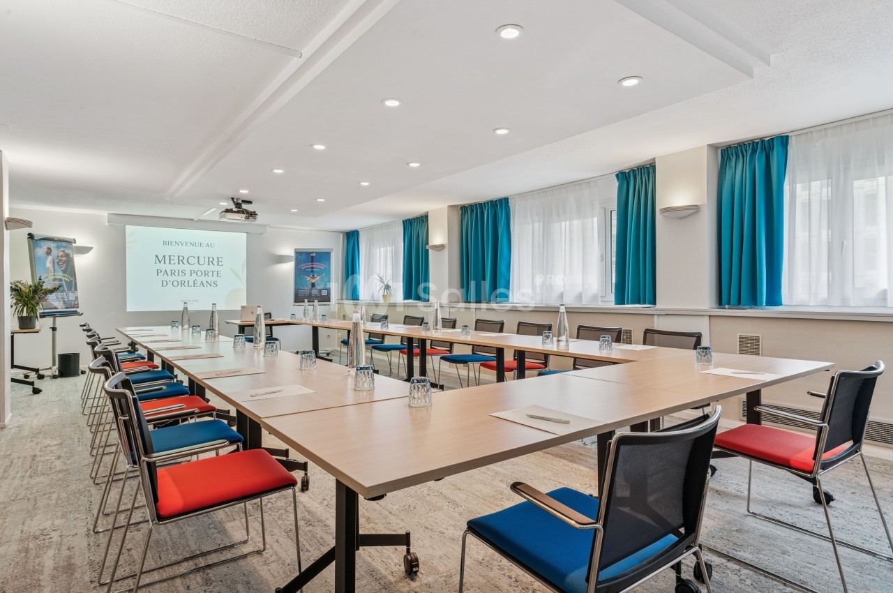 Salle de réunion lumineuse avec tables disposées en U, chaises colorées et écran de projection.