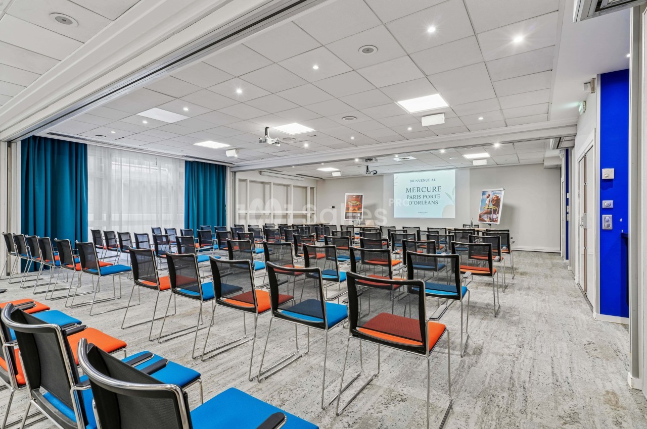 Salle de conférence lumineuse avec des rangées de chaises colorées, écran de projection et rideaux bleus.