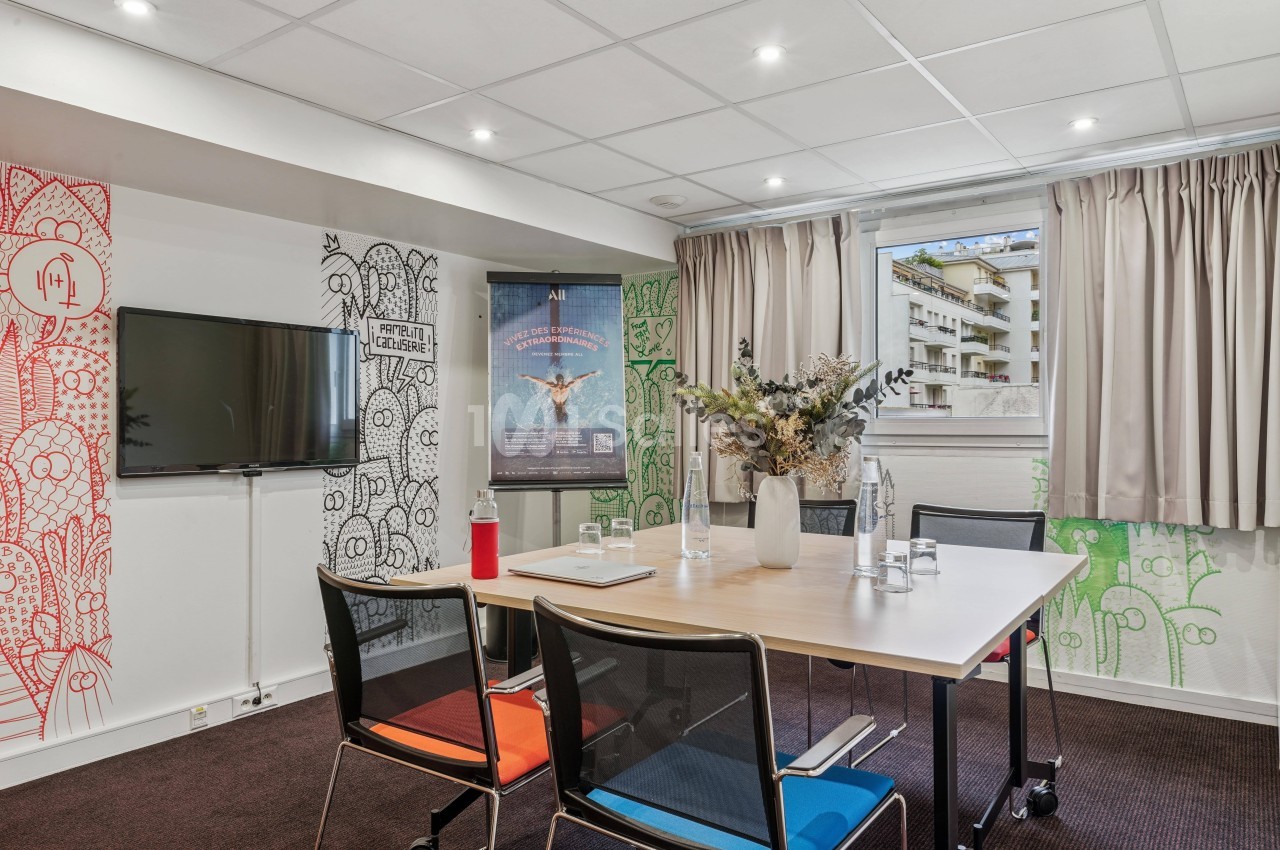 Salle de réunion lumineuse avec table, chaises, écran mural, affiches décoratives et fenêtre donnant sur des immeubles.