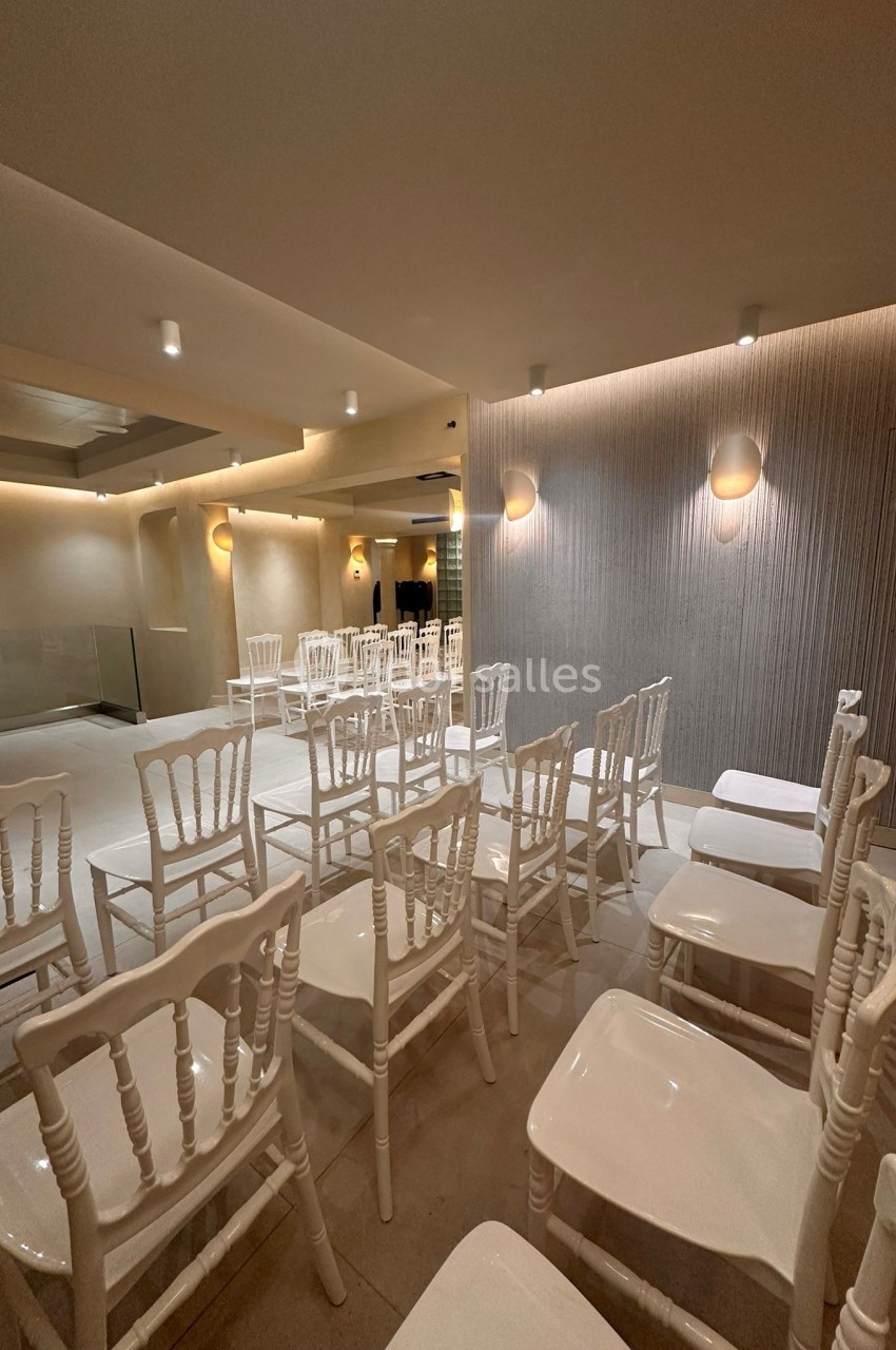 Salle lumineuse avec des chaises blanches alignées, murs texturés et éclairage mural discret.