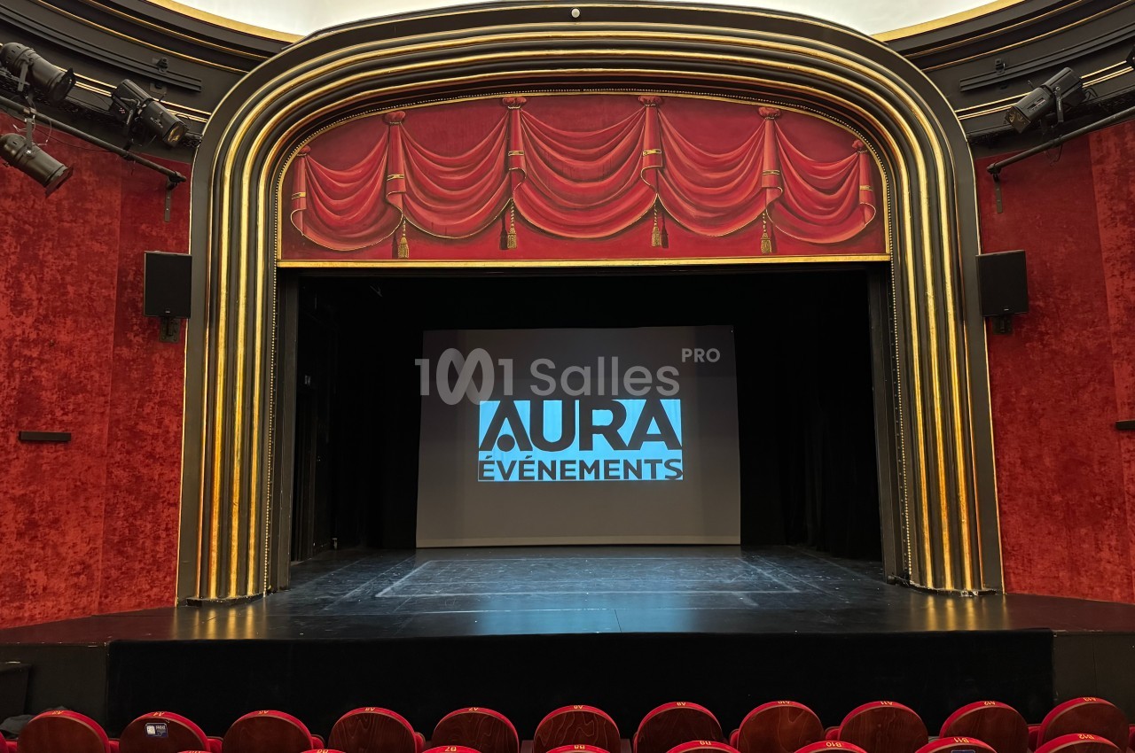 Scène de théâtre avec rideaux rouges, écran central affichant ’Aura Événements’, et rangées de sièges vides au premier plan.