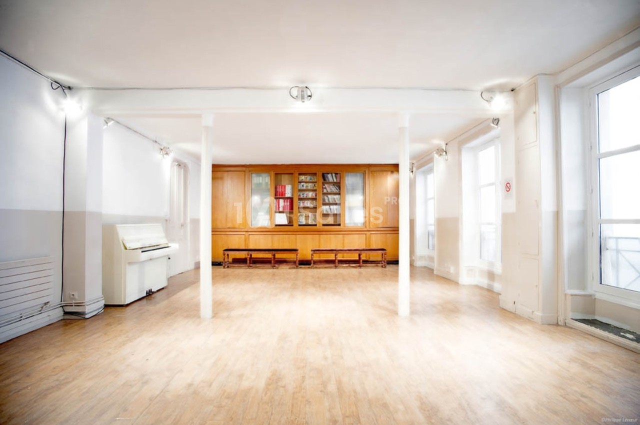 Salle lumineuse avec parquet clair, murs blancs, bibliothèque en bois, piano et grandes fenêtres laissant entrer la lumière.