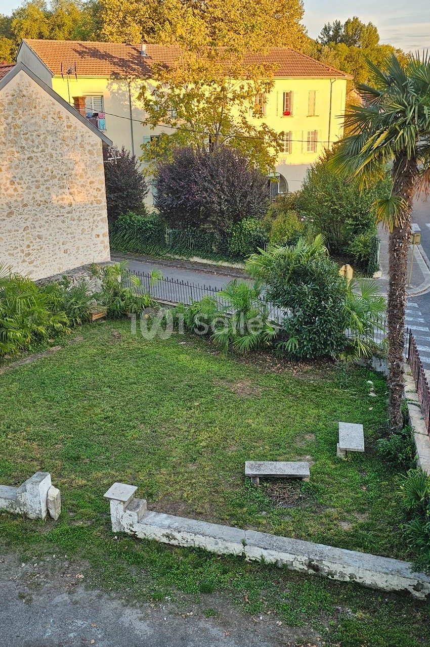 Jardin avec pelouse, bancs en pierre, palmiers et arbres, entouré de bâtiments en pierre et végétation.
