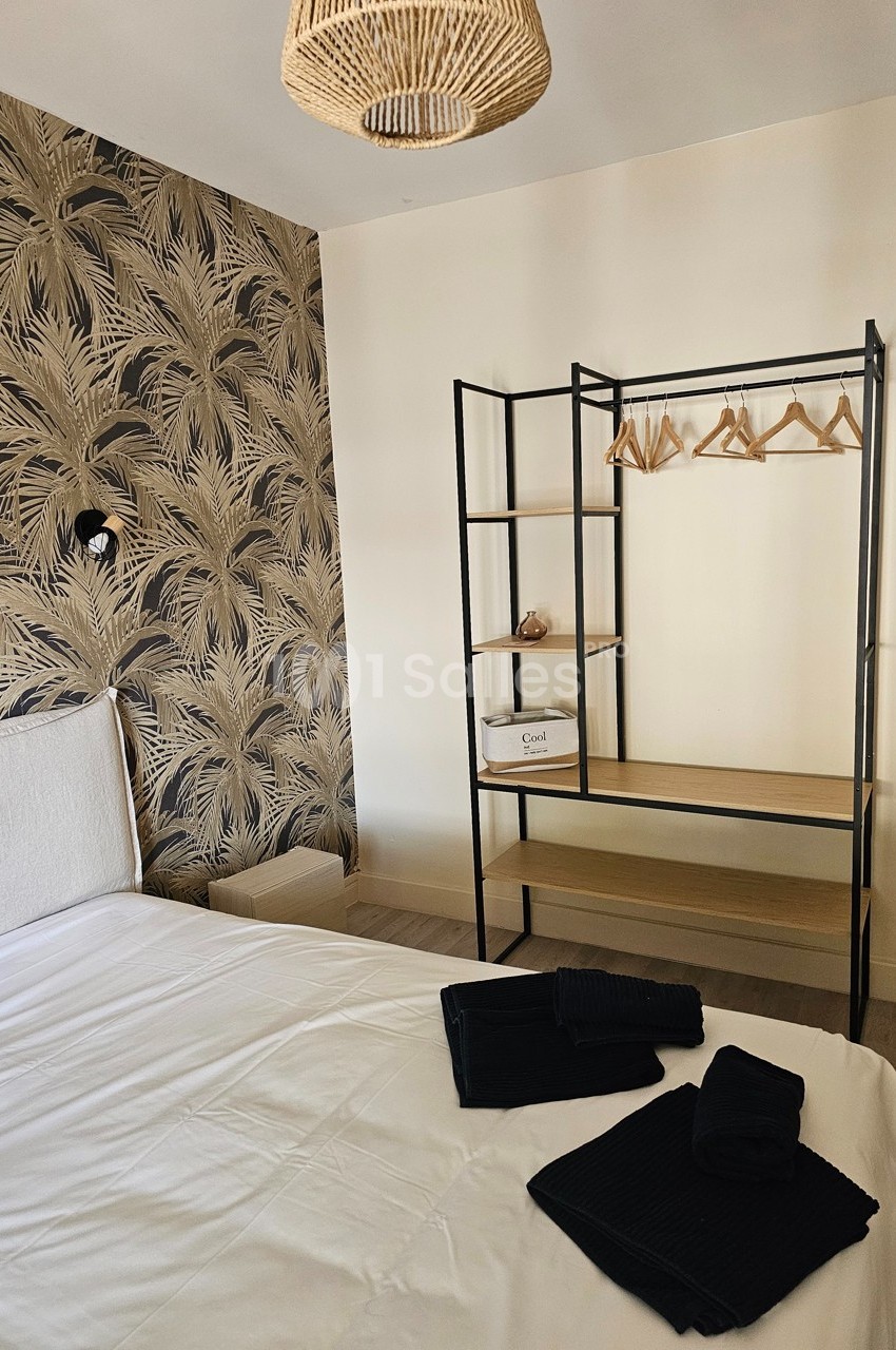 Chambre avec lit blanc, portant en métal noir avec cintres, mur à motif végétal et suspension en rotin.