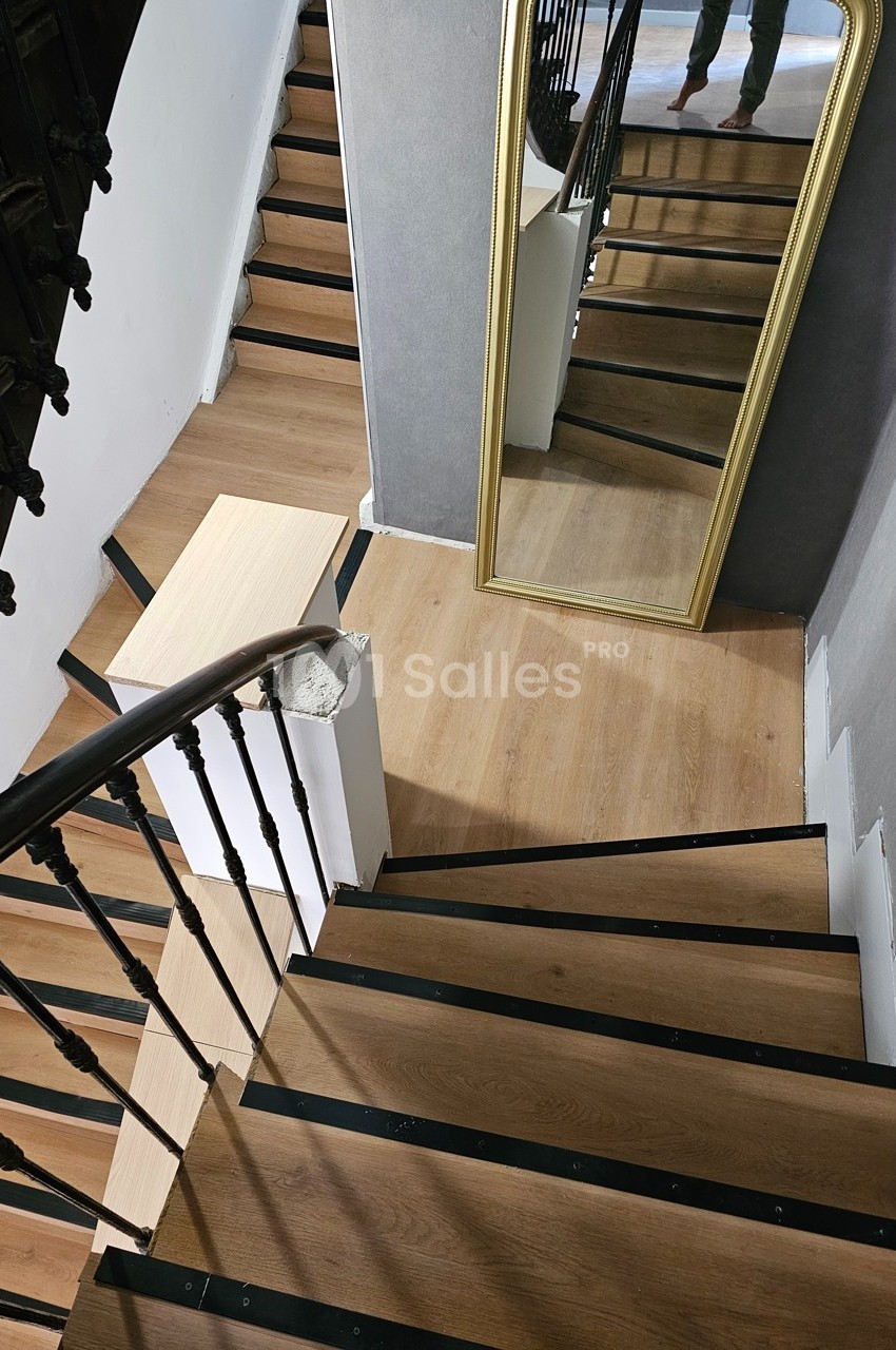Escalier en bois avec rampe en métal noir, grand miroir doré appuyé contre un mur gris.