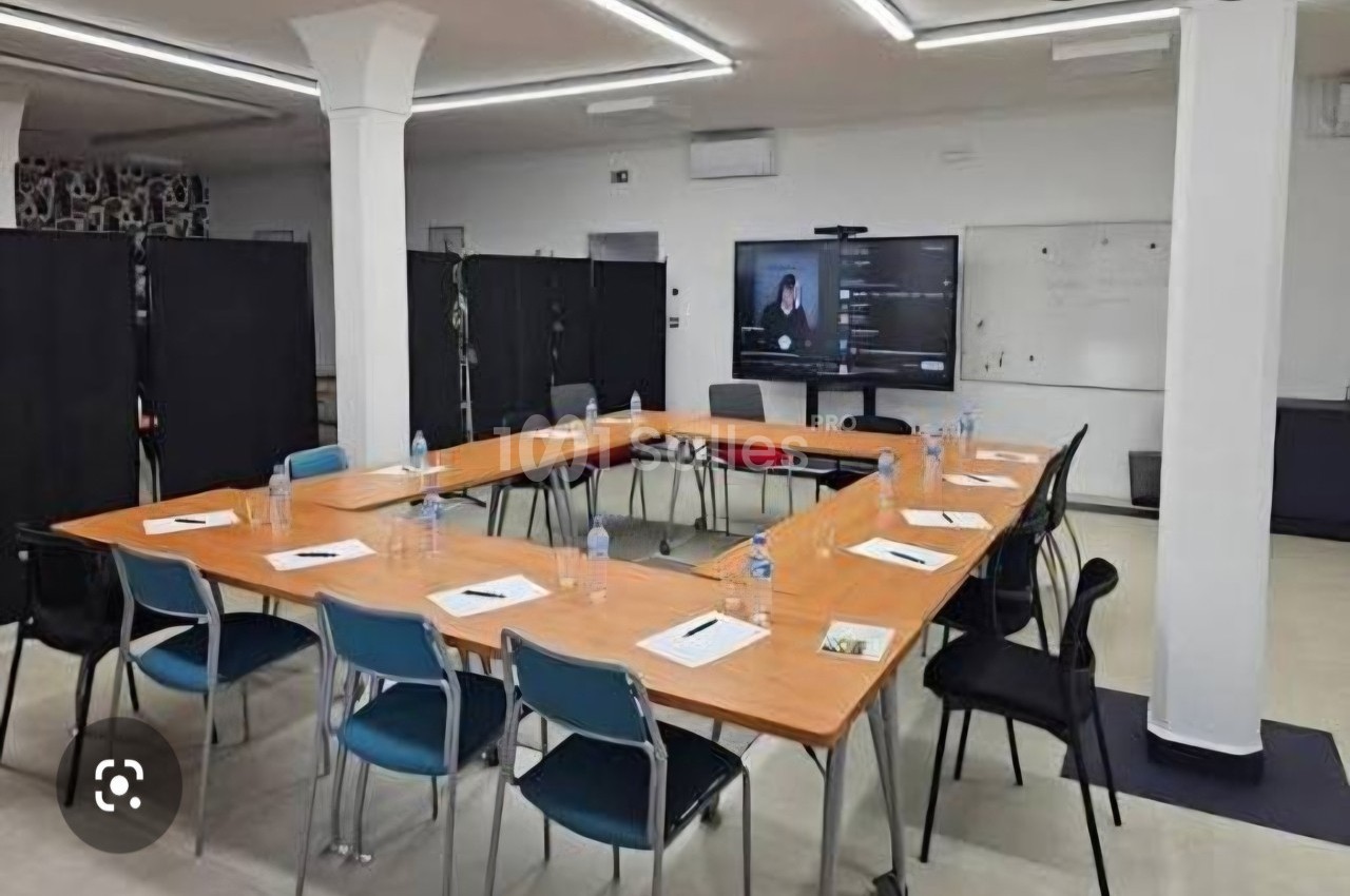 Salle de réunion avec tables disposées en U, chaises, documents, bouteilles d'eau et écran affichant une visioconférence.