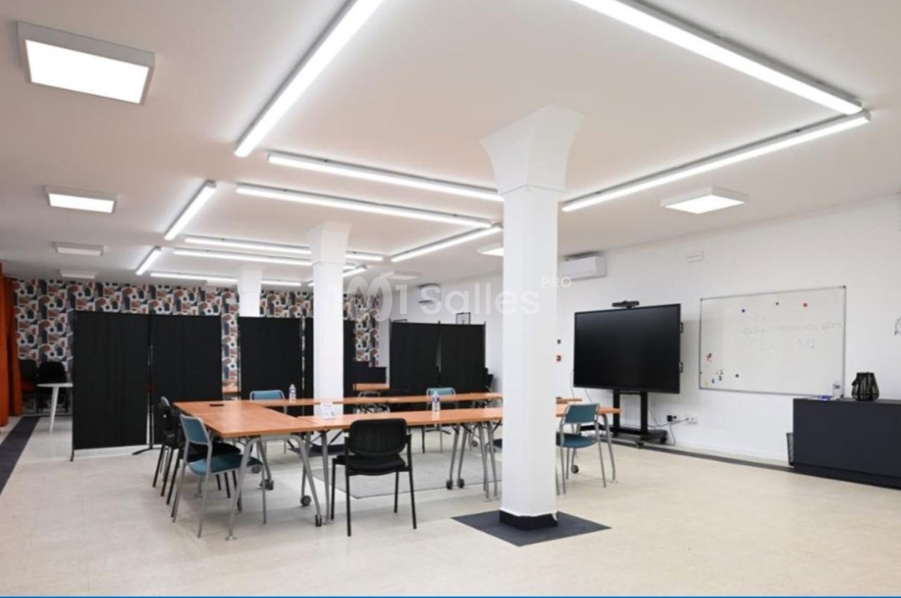 Salle lumineuse avec tables en bois, chaises, tableau blanc, écran et panneaux de séparation noirs.