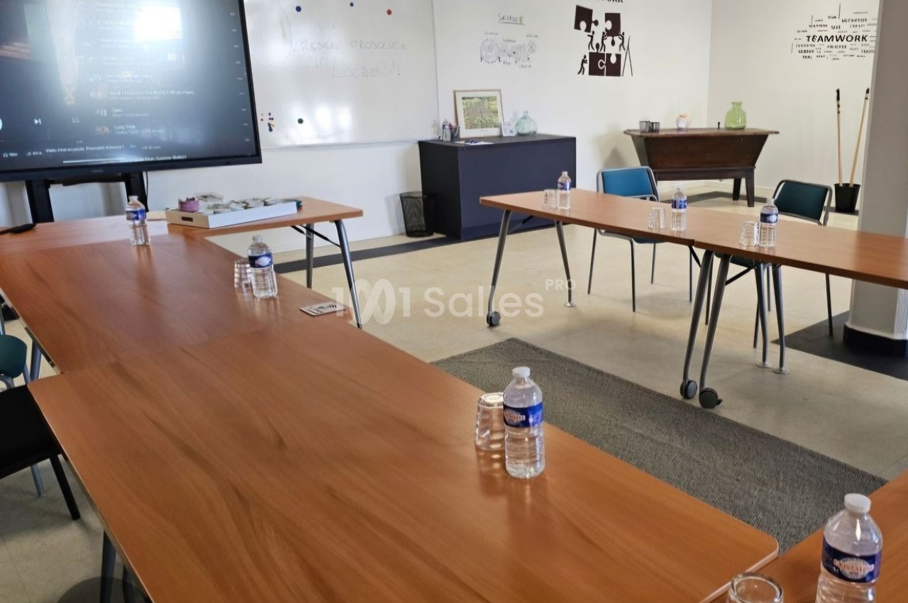 Salle de réunion avec des tables en bois disposées en U, des bouteilles d'eau et un écran de présentation.