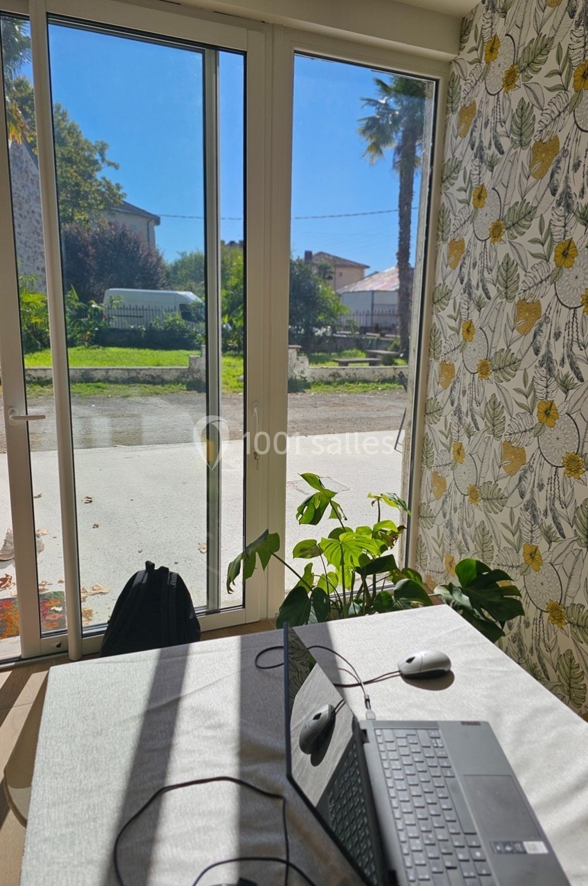 Vue depuis une pièce lumineuse avec une table, un ordinateur portable et une porte-fenêtre donnant sur un jardin ensoleillé.