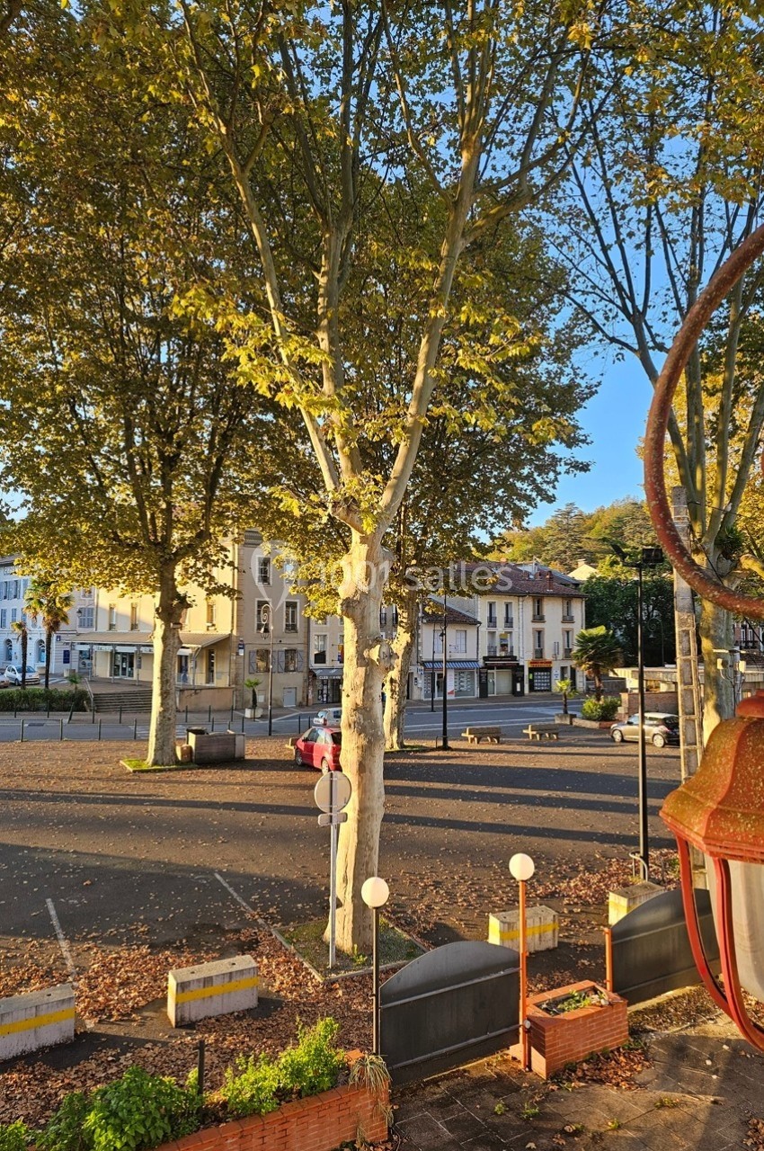 Place bordée d'arbres sous une lumière dorée, avec des bâtiments en arrière-plan et des lampadaires au premier plan.