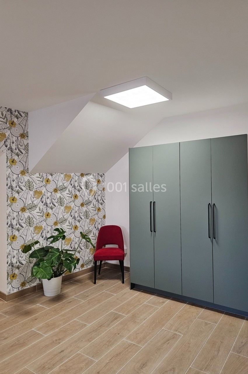Chambre lumineuse avec placard gris, chaise rouge, plante en pot et mur décoré de papier peint floral.