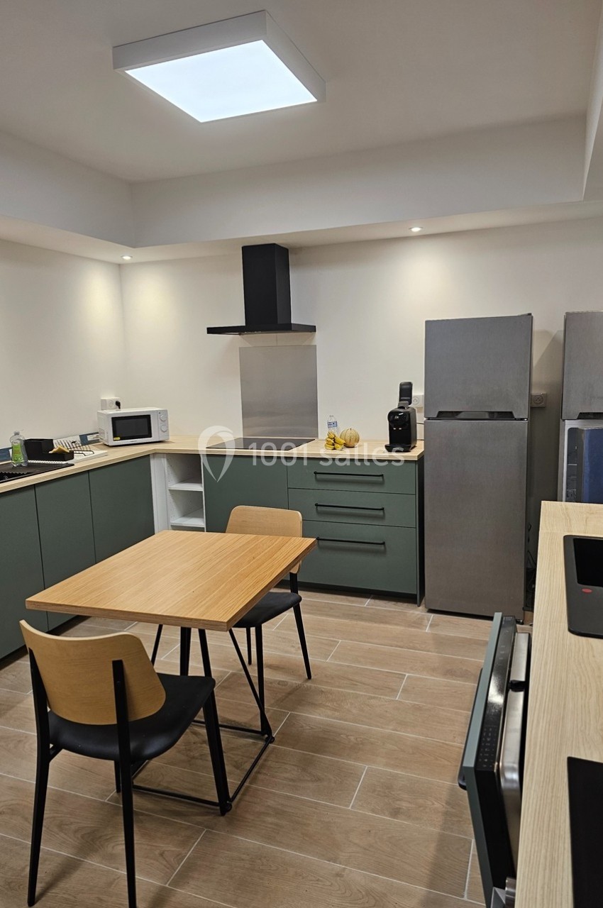 Cuisine moderne avec meubles verts, table en bois, chaises noires, réfrigérateur, micro-ondes et éclairage au plafond.