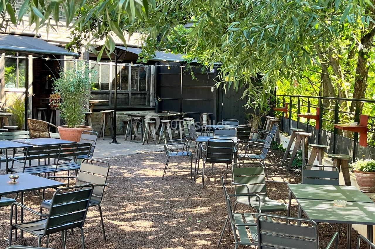 Terrasse extérieure ombragée avec tables et chaises en métal, entourée de végétation et de parasols.