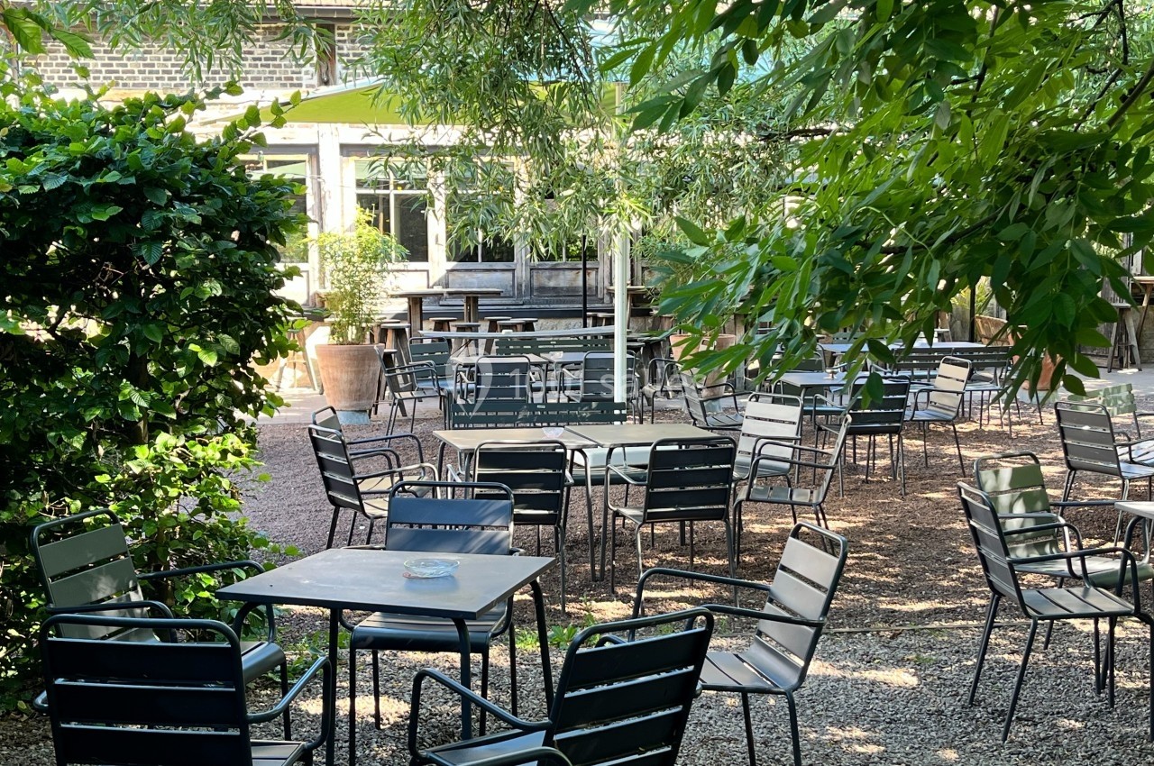 Terrasse extérieure ombragée avec des tables et chaises en métal, entourée de végétation et de lumière naturelle.