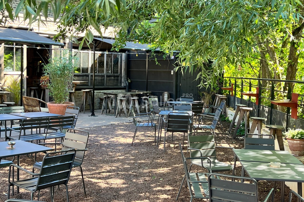 Terrasse extérieure ombragée avec tables et chaises en métal, entourée de végétation et éclairée par la lumière naturelle.