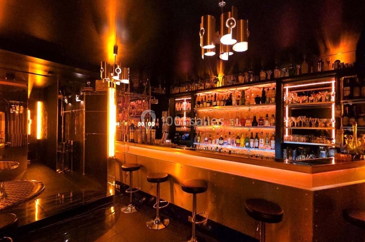 Bar moderne avec éclairage tamisé orange, comptoir en bois, tabourets hauts et étagères remplies de bouteilles.