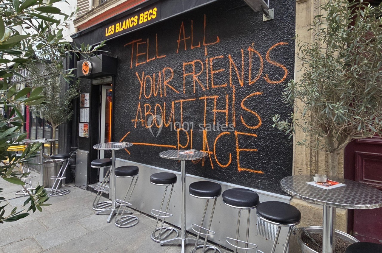 Façade d'un café avec un mur noir portant un message écrit en orange et des tables hautes avec tabourets devant.