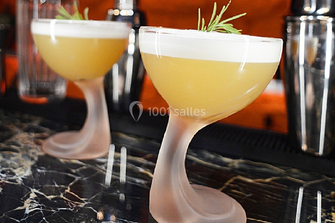 Deux cocktails jaunes dans des verres au pied incurvé, garnis de romarin, posés sur un comptoir en marbre sombre.