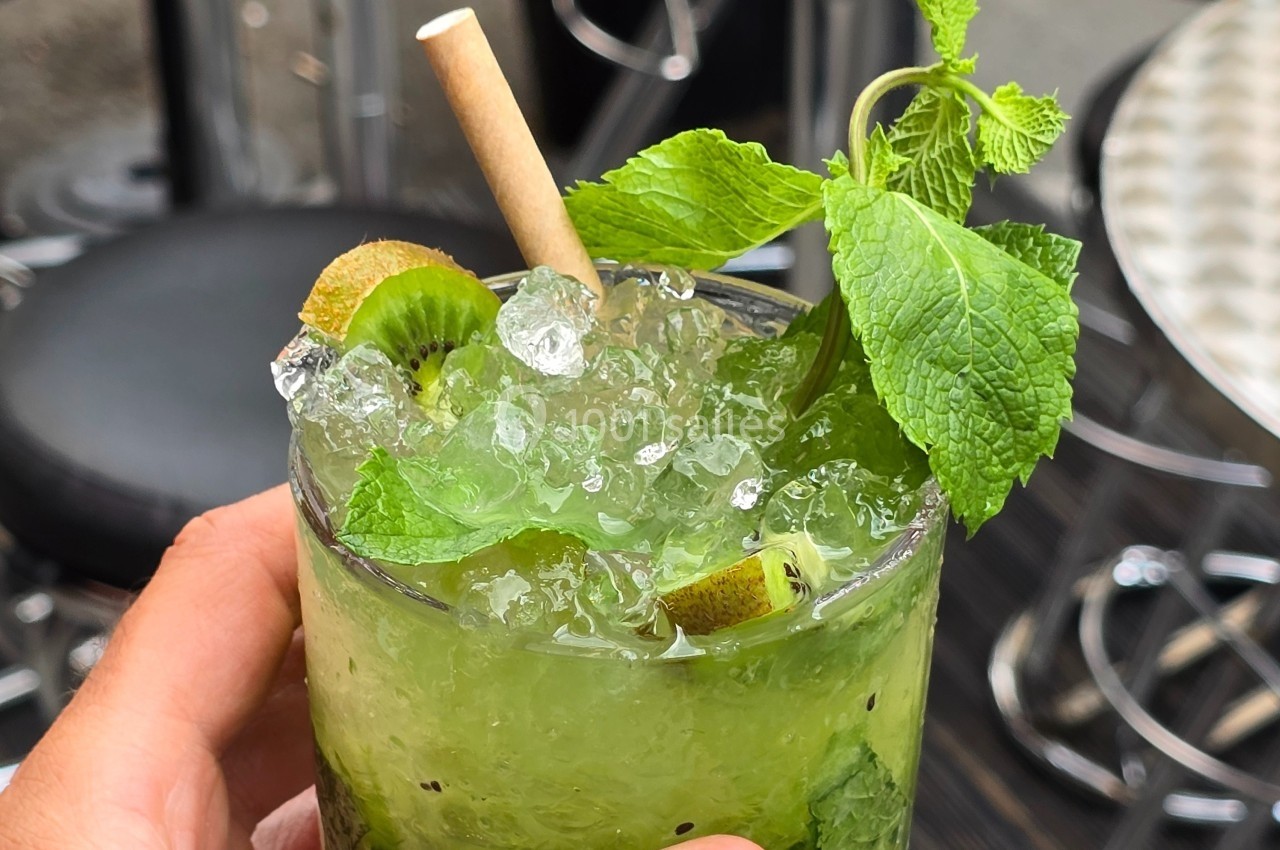 Verre de cocktail vert garni de glace pilée, feuilles de menthe et rondelles de kiwi, tenu par une main.