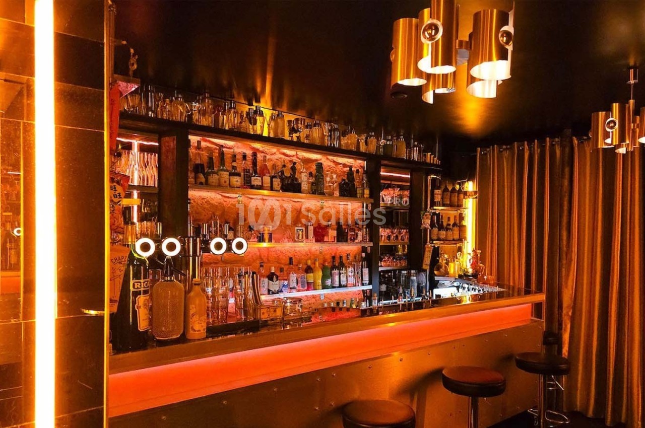 Bar avec étagères illuminées contenant des bouteilles, tabourets noirs et éclairage chaleureux dans une ambiance tamisée.