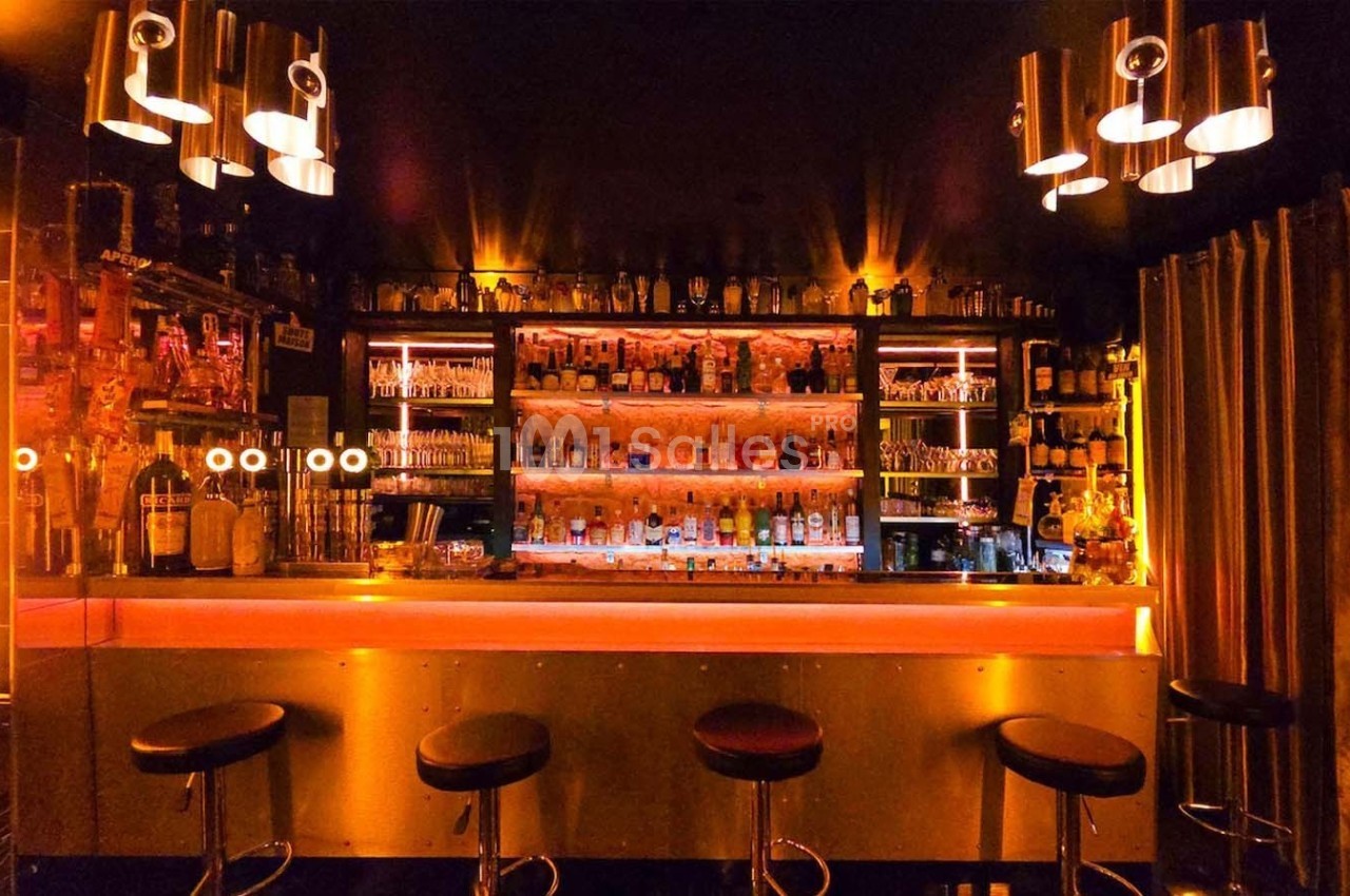 Bar avec comptoir éclairé, tabourets noirs, étagères remplies de bouteilles et ambiance lumineuse tamisée.