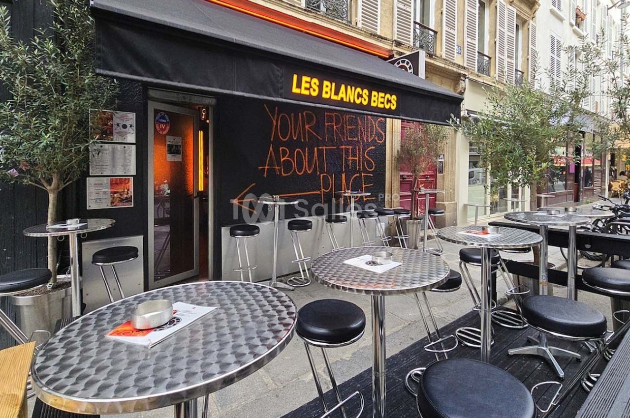 Terrasse d'un café avec tables et chaises en métal, devant une façade noire avec une enseigne lumineuse et un graffiti…