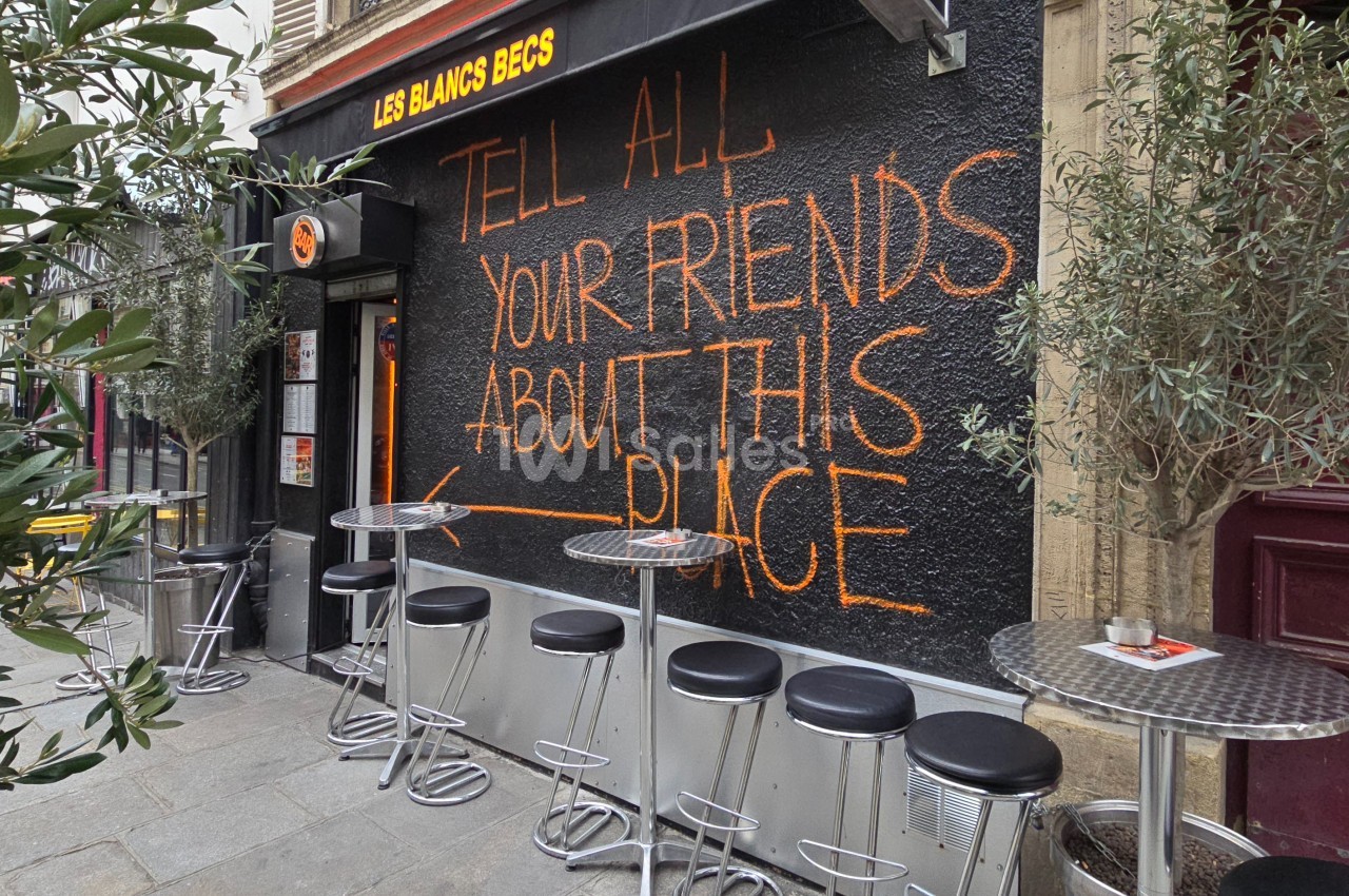 Façade d'un café avec des tables hautes et un mur noir portant une inscription orange manuscrite.