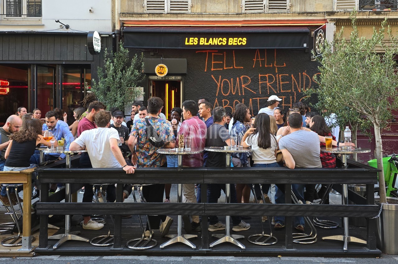 Groupe de personnes attablées en terrasse d'un café animé, avec une enseigne et un mur noir portant des inscriptions.