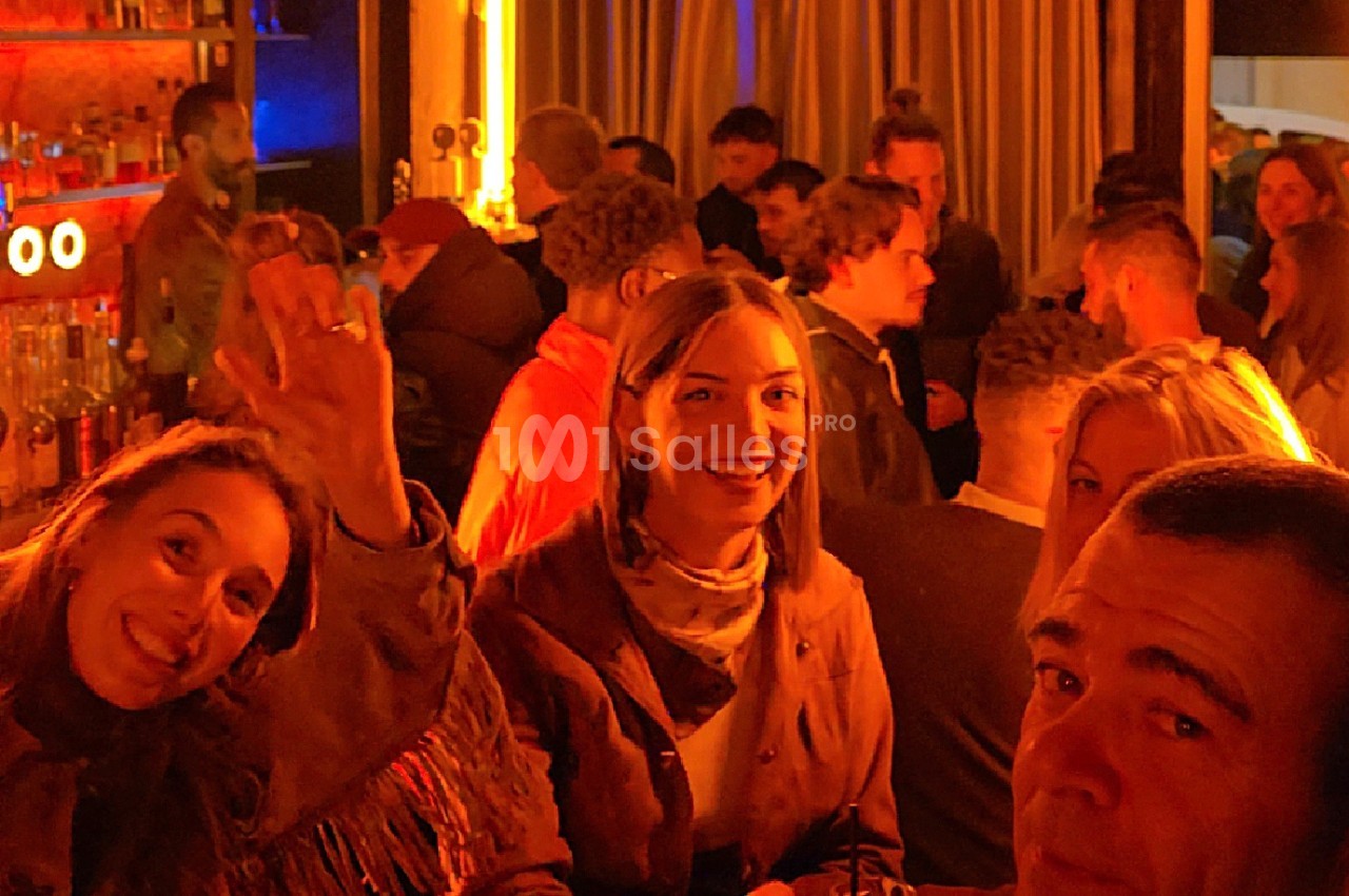 Groupe de personnes souriantes dans un bar animé, éclairé par des lumières chaudes et entouré d'autres clients.