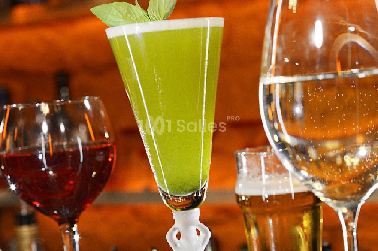 Verres de boissons variées, incluant un cocktail vert garni de feuilles, posés sur un bar éclairé.