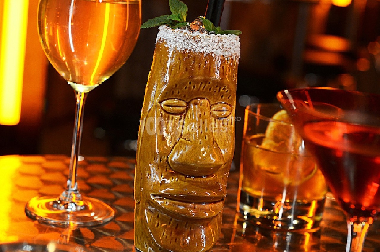Verre tiki sculpté rempli d'un cocktail, entouré de deux autres verres de boissons sur une table dorée.